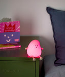 Pink glowing bedtime lamp shaped like a smiling character with small arms and legs, sitting on a green bedside table next to a birthday book.