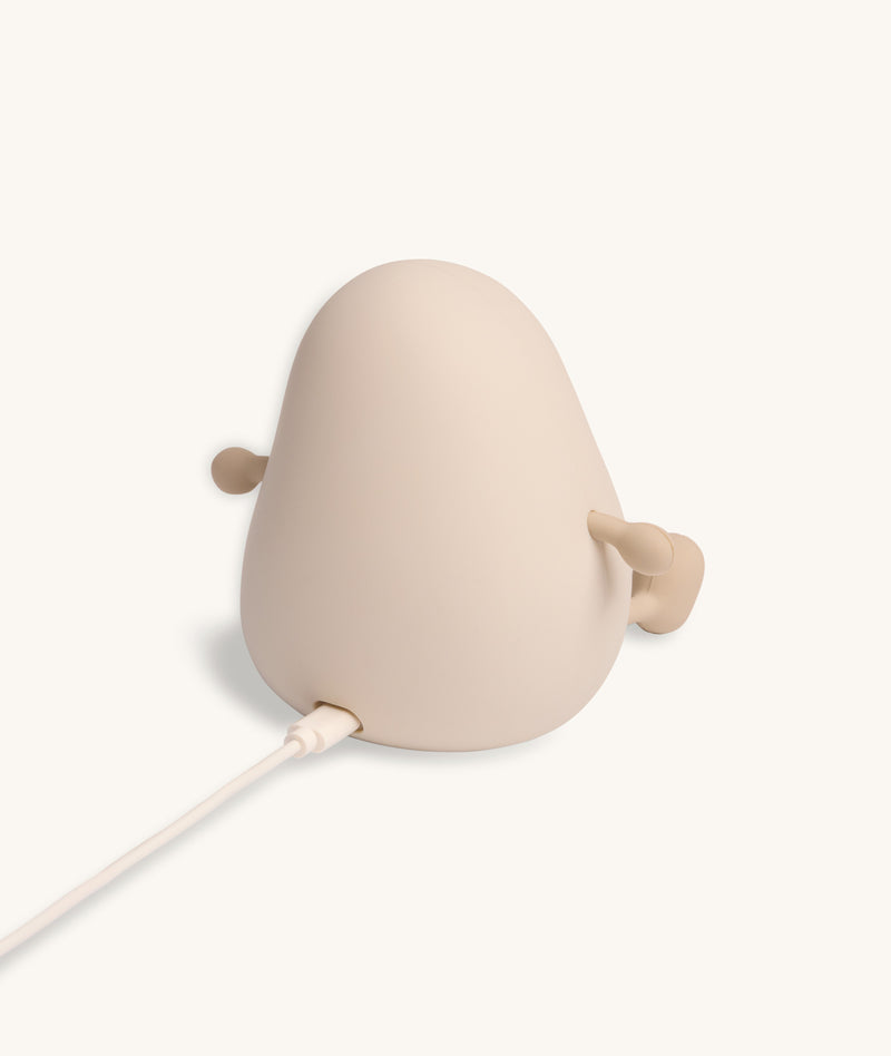 Back view of a soft-touch silicone bedtime lamp shaped like a rounded character with small arms and legs, shown plugged in with a charging cable against a plain light background.