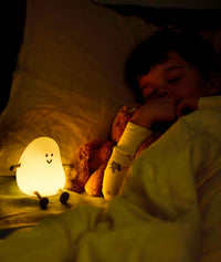 Child sleeping in bed cuddling a stuffed toy, with a softly glowing yellow bedtime lamp shaped like a smiling character placed nearby, casting a warm light in the dark room.