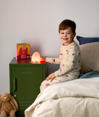 Smiling child sitting on a bed, touching a glowing orange bedtime lamp shaped like a happy character on a green bedside table.