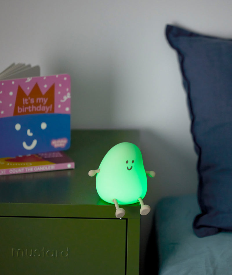Green glowing bedtime lamp shaped like a smiling character with small arms and legs, sitting on a green bedside table next to a childrens birthday book.