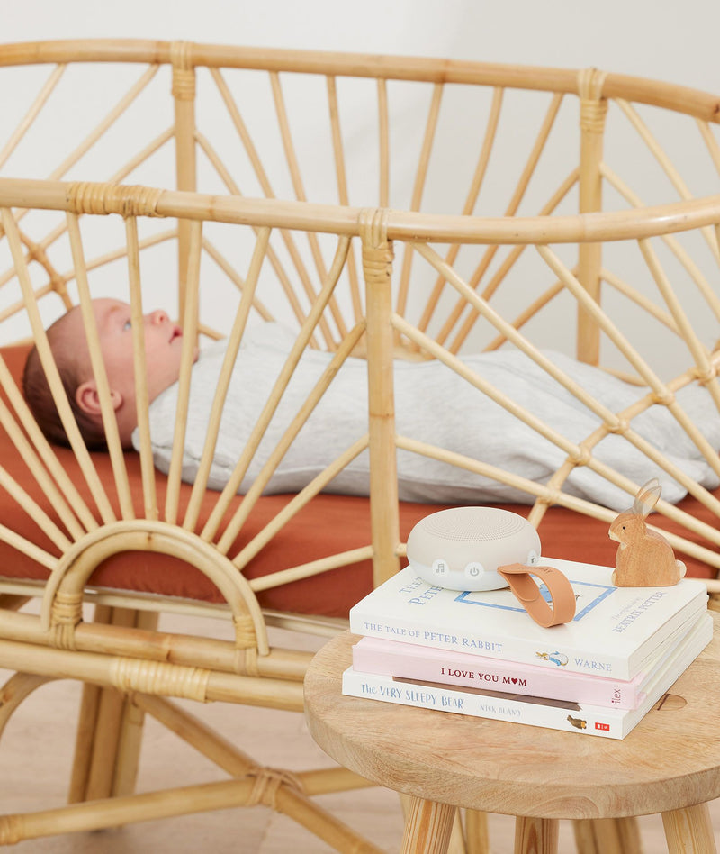 A beige white noise machine with a tan leather strap rests on a stack of children's books next to a baby sleeping in a natural rattan crib.