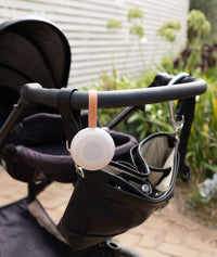 A compact white noise machine by from ergoPouch with a circular speaker design is attached to a pram handle using a tan leather strap embossed with the name “Drift Away.”