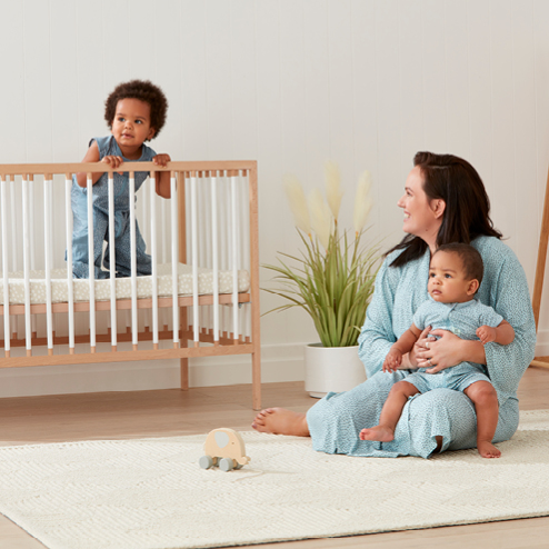 Mom sitting on ground holding baby, looking at toddler in crib