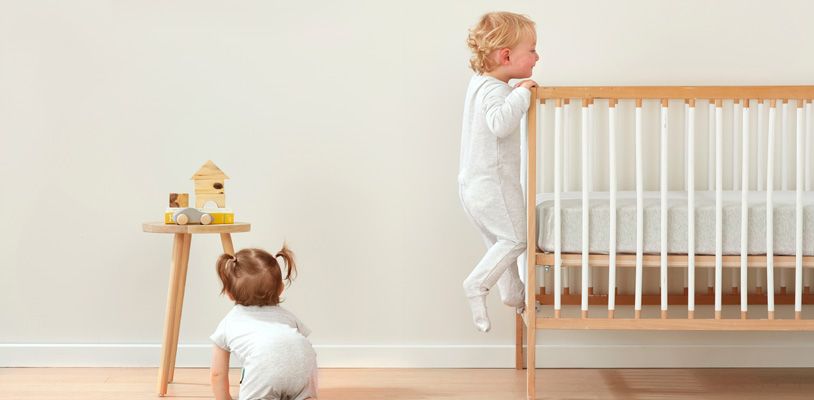 Toddler crawling out of crib while baby crawls on the floor nearby