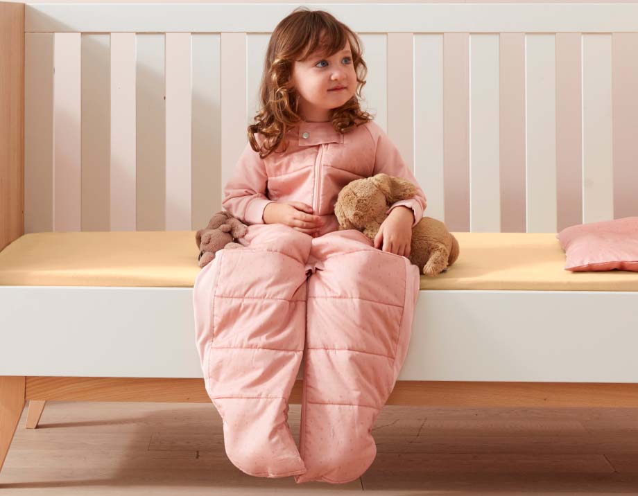 Toddler sitting on edge of bed wearing Sleep Suit Sack in Berries, holding a teddy